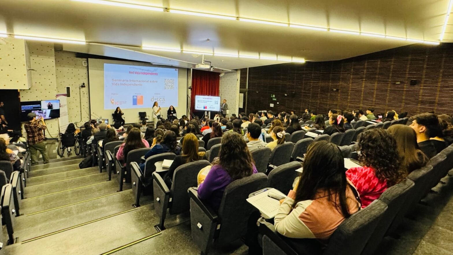 La imagen captura una vista desde el escenario de un auditorio concurrido . Se observa a un orador de pie en el centro, dirigiéndose a una numerosa audiencia sentada en filas de asientos oscuros. A la izquierda, parte de la pantalla de proyección visible menciona el "Seminario Internacional sobre Vida Independiente" y se ven dos panelistas sentados en la parte delantera (uno de ellos en silla de ruedas), con una mesa de conferencias y micrófonos en primer plano a la derecha.