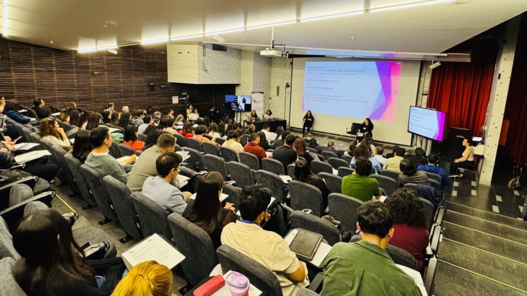 La fotografía muestra una vista desde las últimas filas de un auditorio repleto de asistentes sentados en asientos acolchados grises. Al fondo, en el escenario, se aprecian dos oradoras o panelistas de espaldas al público, interactuando frente a una pantalla de proyección que muestra texto relacionado con "MARCO POLÍTICO" y la Convención sobre los Derechos de las Personas con Discapacidad. El ambiente es el de un evento educativo o profesional bien atendido.