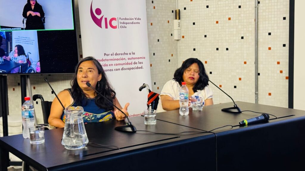 La fotografía muestra un primer plano de dos mujeres sentadas en una mesa de panel durante una conferencia. La mujer de la izquierda está hablando activamente con un micrófono cerca, mientras la de la derecha escucha. Detrás de ellas, un banner grande muestra el logo de "Fundación Vida Independiente Chile" y el lema "Por el derecho a la autodeterminación, autonomía y vida en comunidad de las personas con discapacidad".