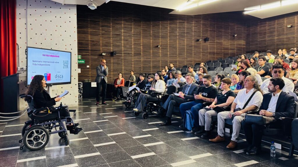 La fotografía capta una sesión de panel o conferencia en un auditorio. Una mujer en silla de ruedas se encuentra en primer plano a la izquierda, hablando o leyendo desde unas notas. Detrás de ella, una pantalla de proyección muestra el título "Seminario Internacional sobre Vida Independiente". En el centro ya la derecha, se observa a un panel de invitados sentados en una primera fila, junto a otros asistentes en las filas posteriores.