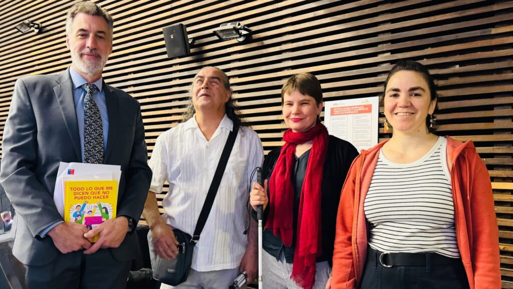 La fotografía es un plano medio de cuatro personas (dos hombres y dos mujeres) posando juntas, probablemente participantes o expositores del evento. El hombre de la izquierda sostiene un libro o folleto de colores vivos , y la mujer a su derecha usa un bastón blanco y una bufanda roja. Todos posan sonriendo o mirando a la cámara, con una pared de tablones de madera horizontales como fondo.